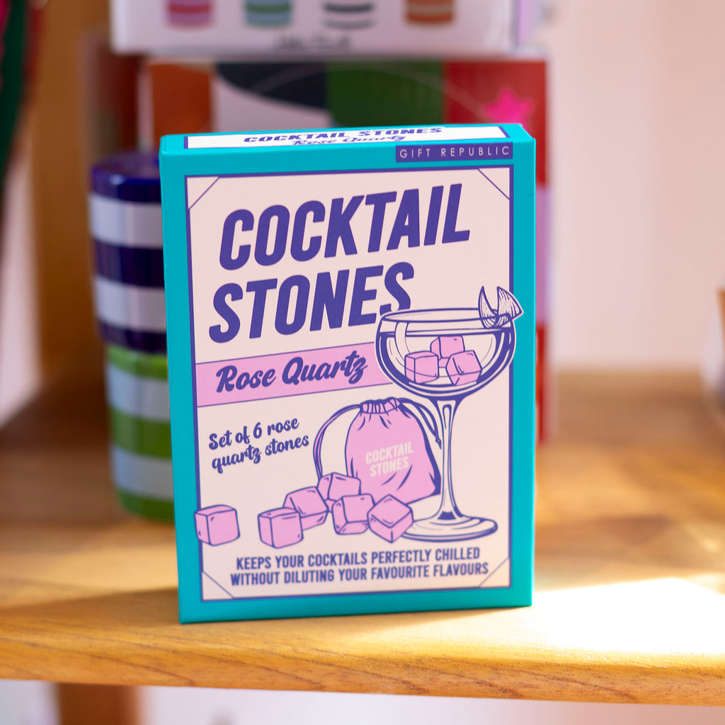 A boxed set of Cocktail Stones by Gift Republic, specifically the Rose Quartz flavor, standing on a wooden shelf. The packaging features a bright turquoise border with purple and white graphics showing a cocktail glass, scattered stones, and a drawstring bag.