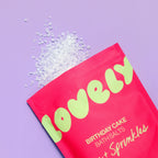 Birthday Cake Bath Salts in bright pink packaging with mineral bath salts scattered on a lilac background.
