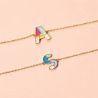 Colourful enamel A and S initial bracelets with delicate gold-tone chains displayed on a pale pink background.