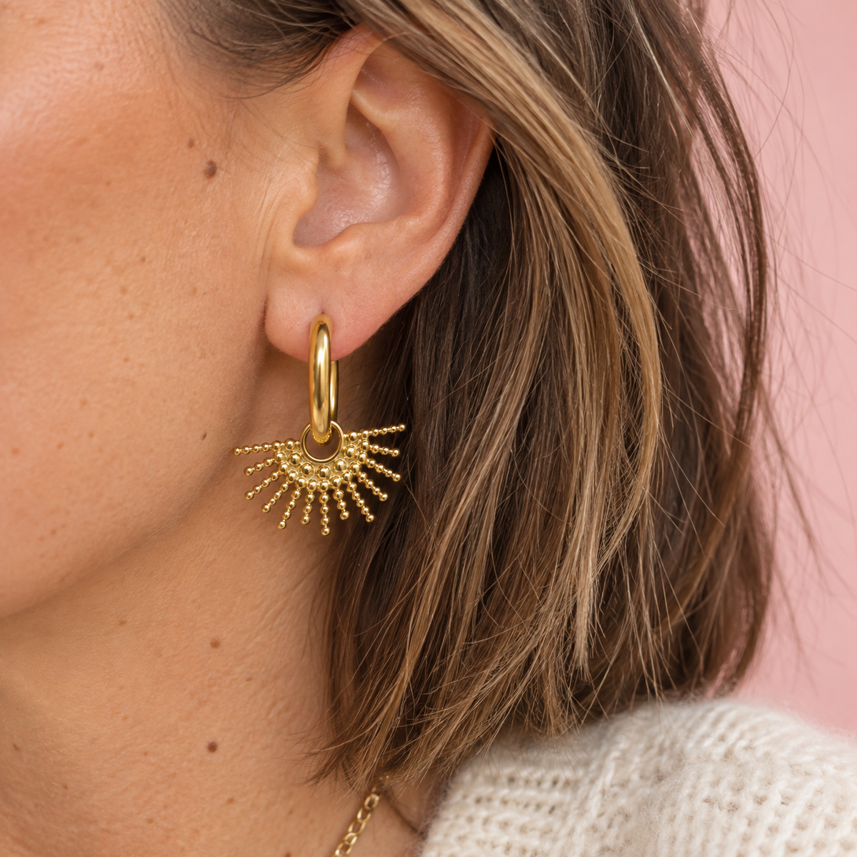 Close up of a woman wearing a gold oval hoop earring with a textured sunburst drop.