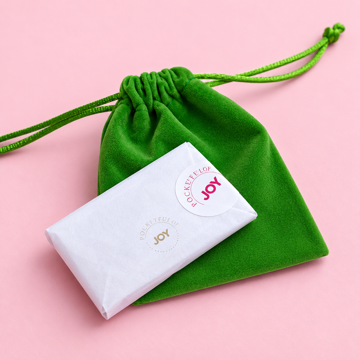 A white wrapped jewellery package with a round white sticker reading 'JOY' and 'VIBRANTFULNESS' lies on top of a green velvet drawstring pouch against a solid pink background.