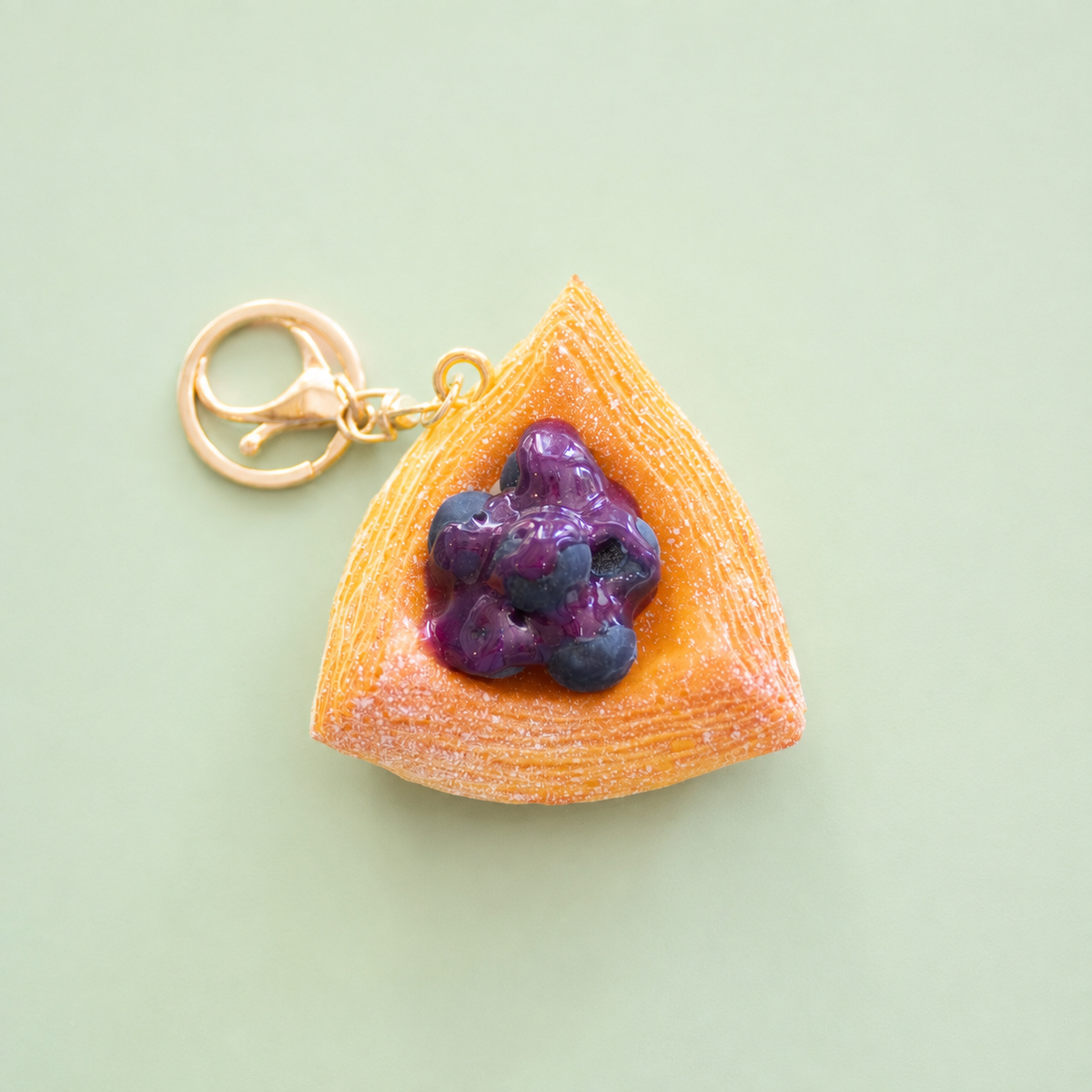 Blueberry Danish pastry bag charm keyring with golden pastry layers, glossy purple blueberries and gold clip on a pale green background.