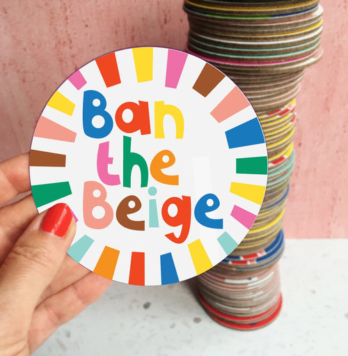 Round white Ban The Beige coaster with colourful lettering and bright striped border, held against a pink background.