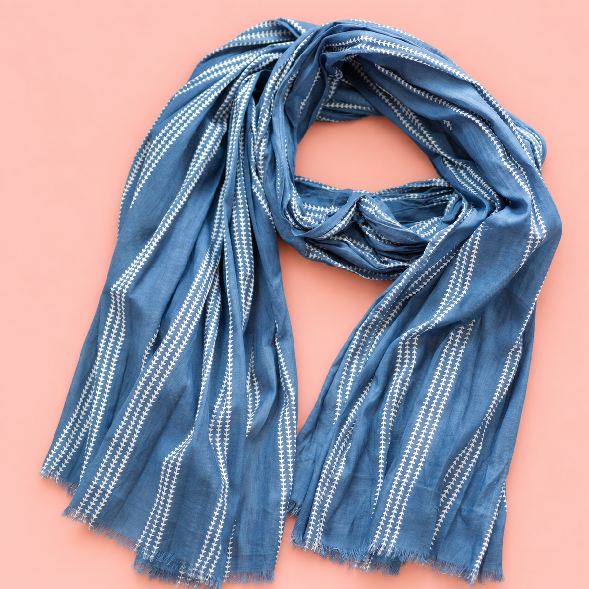 Lightweight blue scarf with white textured stripe detail and softly frayed edges on a peachy pink background.