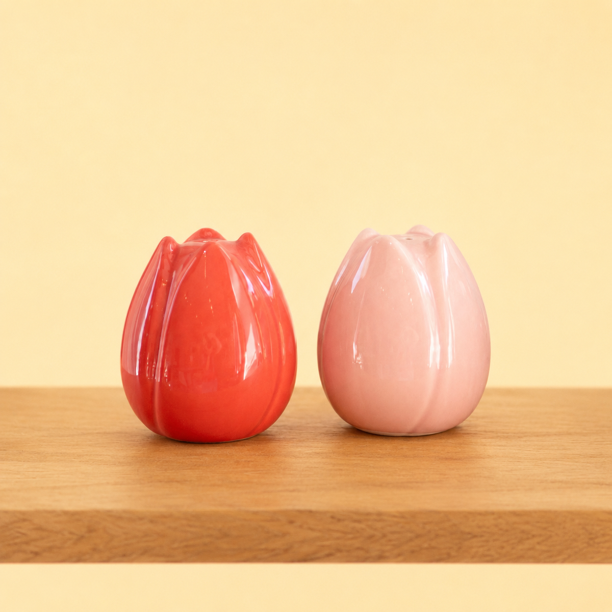 Red and soft pink ceramic tulip shaped salt and pepper shakers displayed on a wooden surface against a pale yellow background.