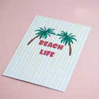 Colourful Beach Life A5 palm tree print with yellow stripes, photographed at an angle against a soft pink background.