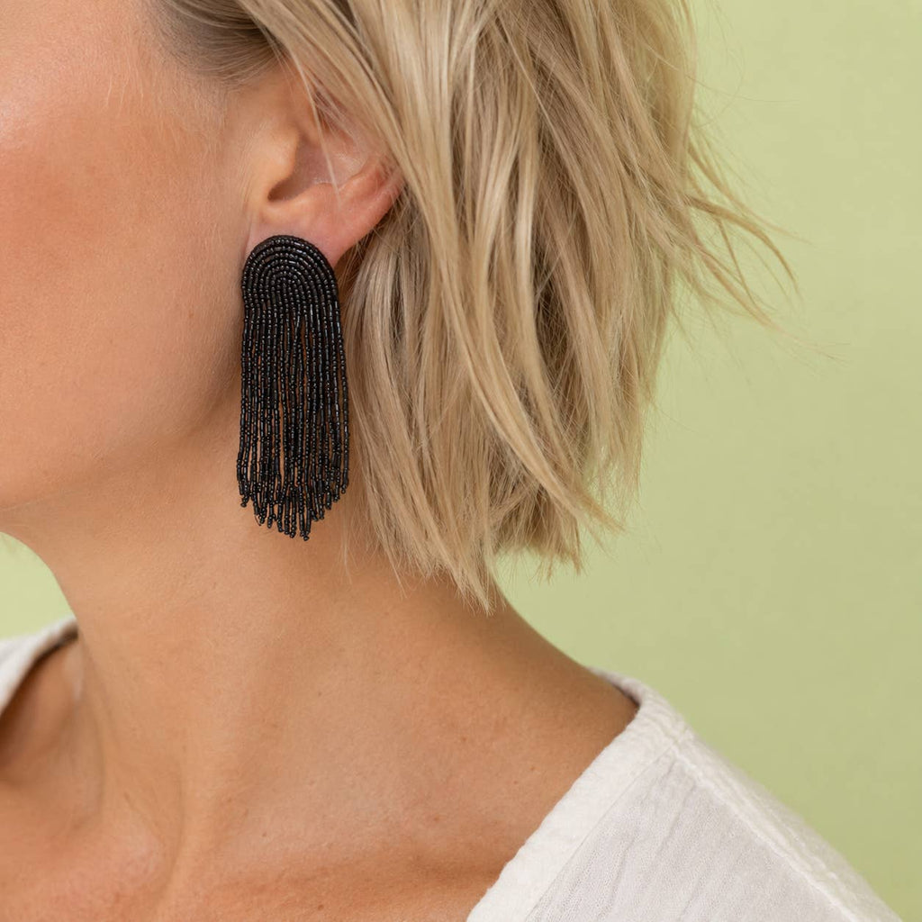 Black beaded fringe statement earring with long cascading strands, styled on the ear against a soft green background.