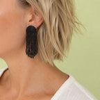 Black beaded fringe statement earring with long cascading strands, styled on the ear against a soft green background.