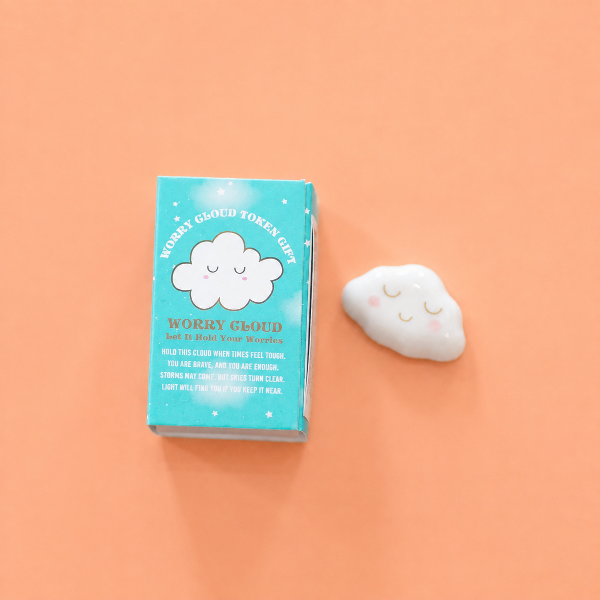 White ceramic Worry Cloud token with smiling face beside its illustrated gift box on a pink background. Available in two designs, selected at random.