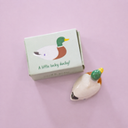 Boxed ceramic lucky duck token with green head, brown chest and white body on a pale pink background.