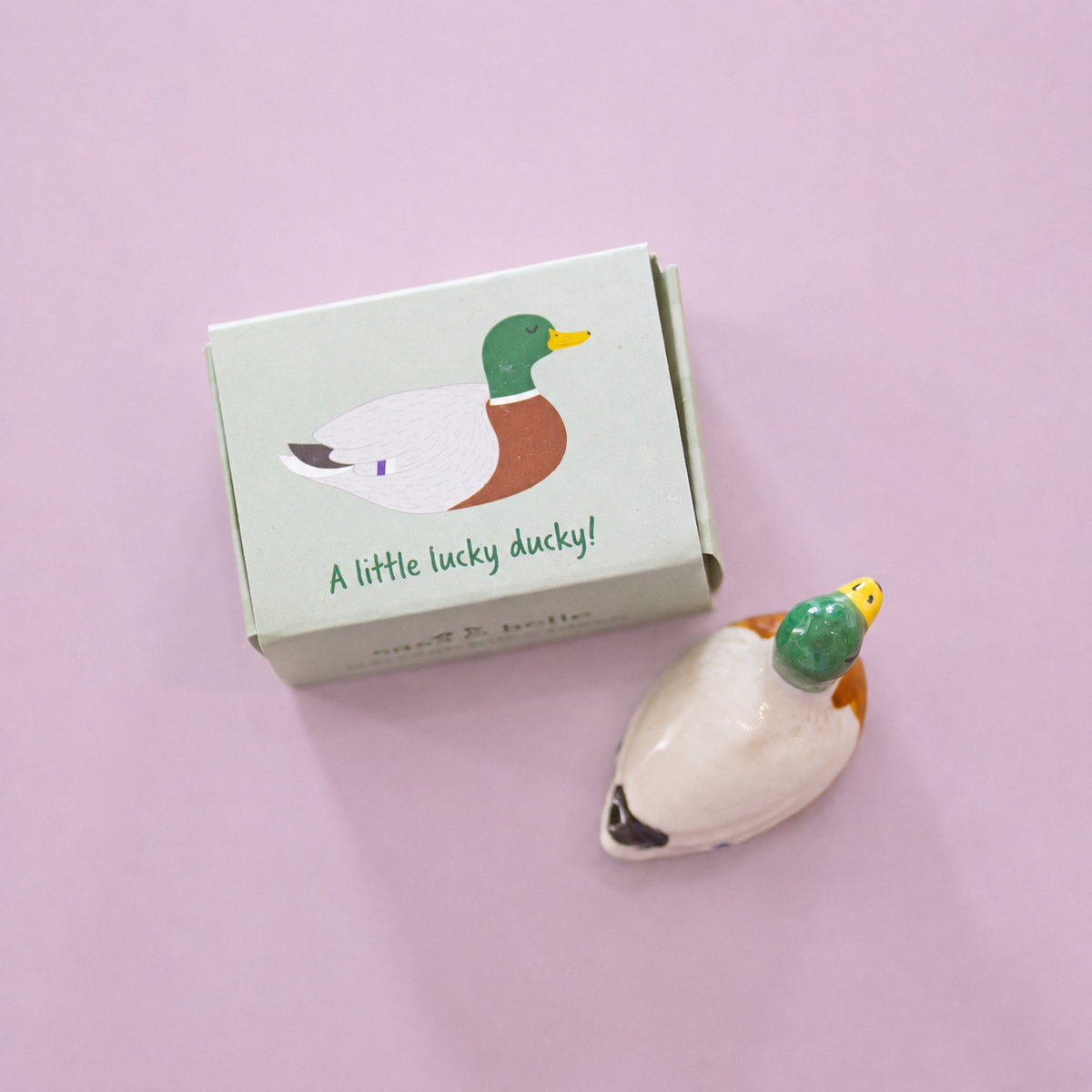 Boxed ceramic lucky duck token with green head, brown chest and white body on a pale pink background.
