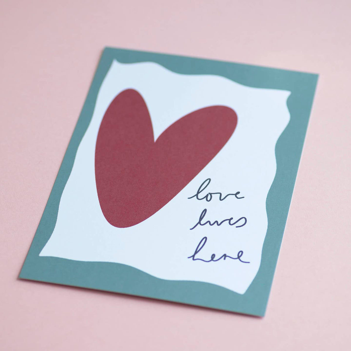 Cheerful Love Lives Here art print featuring a large red heart and handwritten message, styled against a pale pink background.
