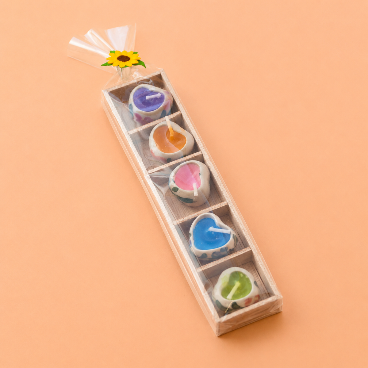 A set of five heart-shaped tea light candles in a clear plastic gift box with a wooden interior, tied with a clear cellophane sleeve and decorated with a small sunflower at the top. The candles are shown in various colors: purple, orange, pink, blue, and green, set against a solid peach background.