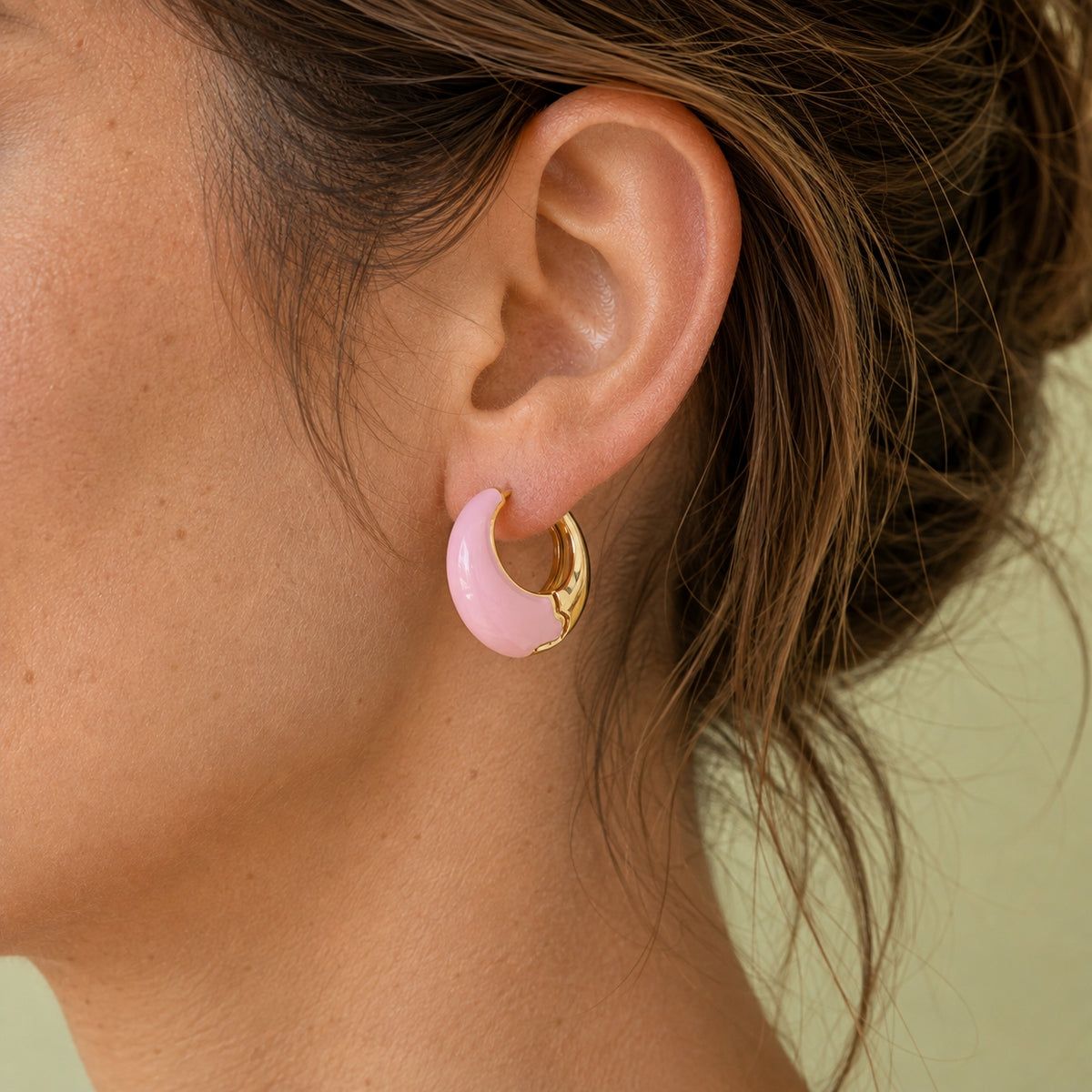 Pale pink and gold-tone enamel huggie hoop earring styled on the ear, showing its chunky curved shape.