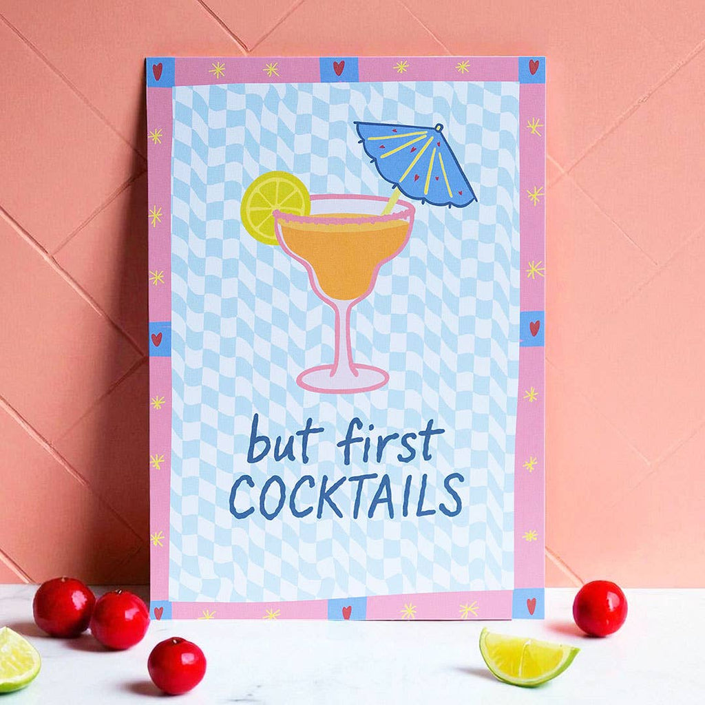 Colourful But First Cocktails A5 print with cocktail, lime and parasol illustrations, styled against a warm coral wall.
