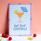 Colourful But First Cocktails A5 print with cocktail, lime and parasol illustrations, styled against a warm coral wall.