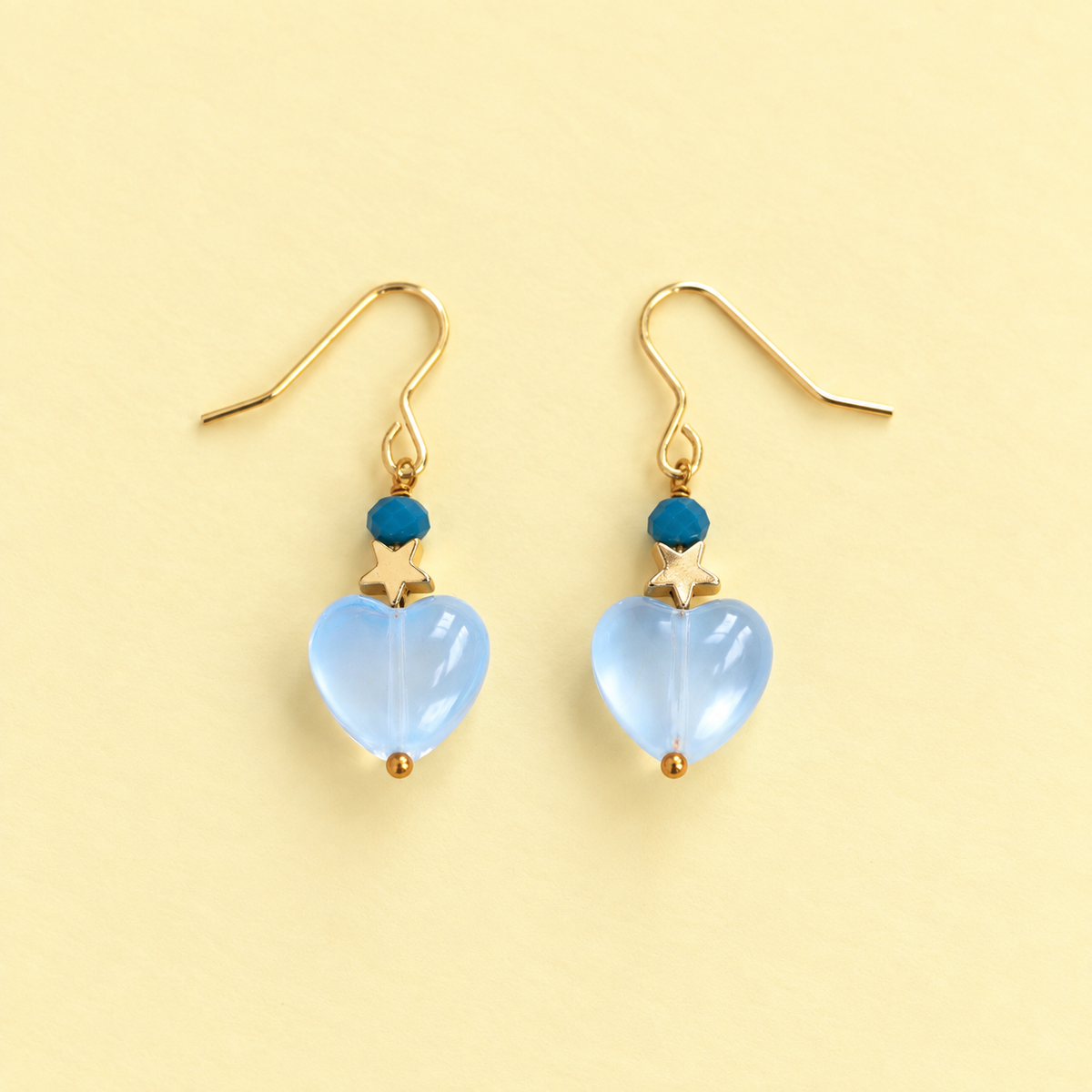 One of a kind gold plated drop earrings with pale blue glass heart beads, tiny gold stars and dark blue faceted beads on a pale yellow background.
