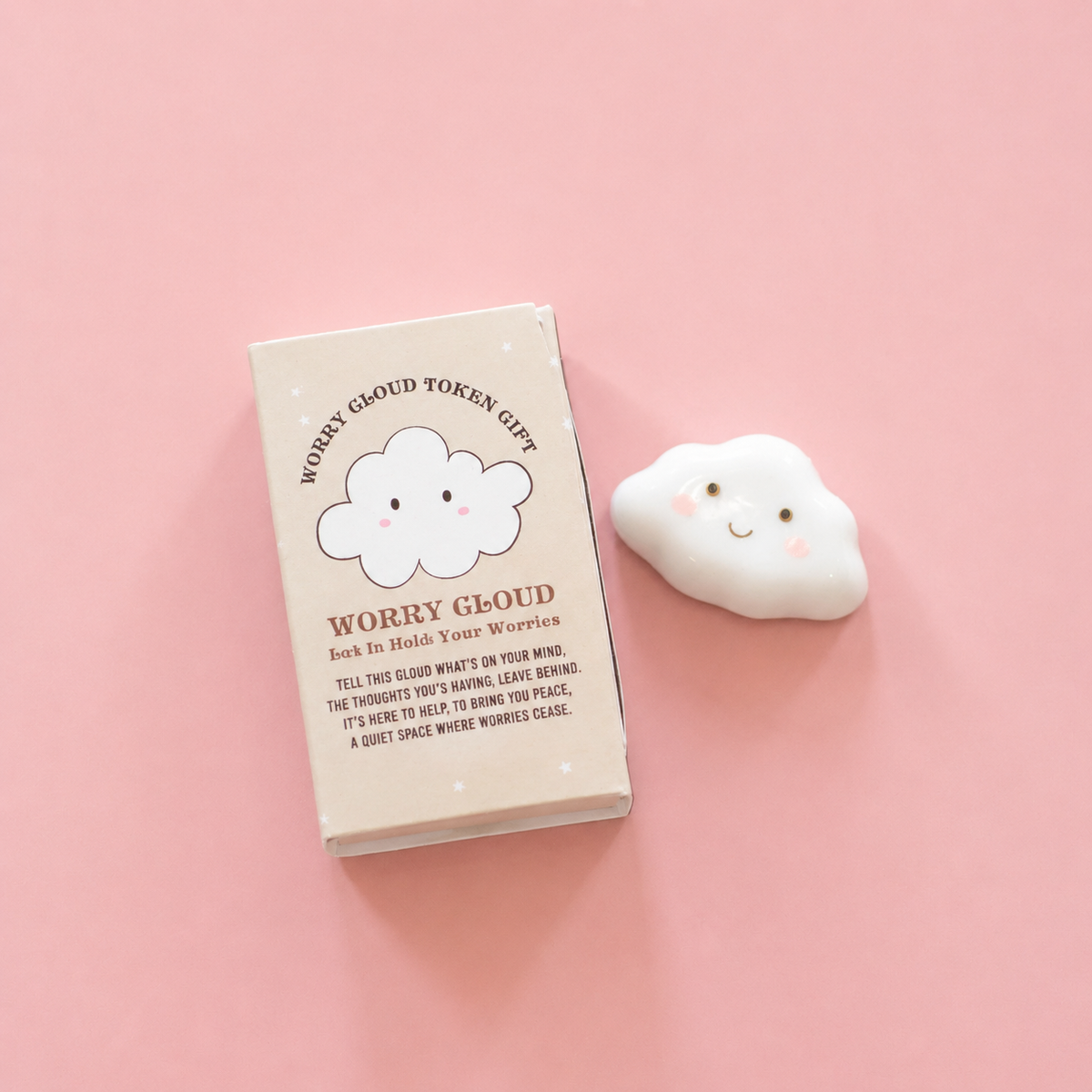 White ceramic Worry Cloud token with smiling face beside its illustrated gift box on a pink background. Available in two designs, selected at random.