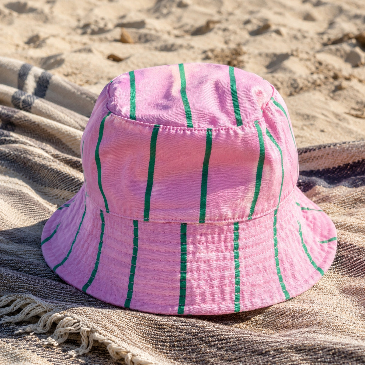Pink and green striped bucket hat displayed on a woven blanket at the beach.