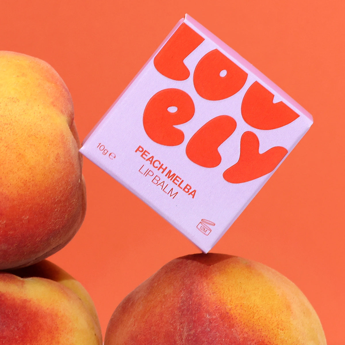Colourful Peach Melba Lip Balm box displayed amongst fresh peaches on an orange