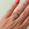 Multicolour enamel gold tone signet ring with sparkling crystal centre worn on a hand.