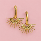 Gold oval hoop earrings with textured beaded sunburst drops on a soft dusky pink background.
