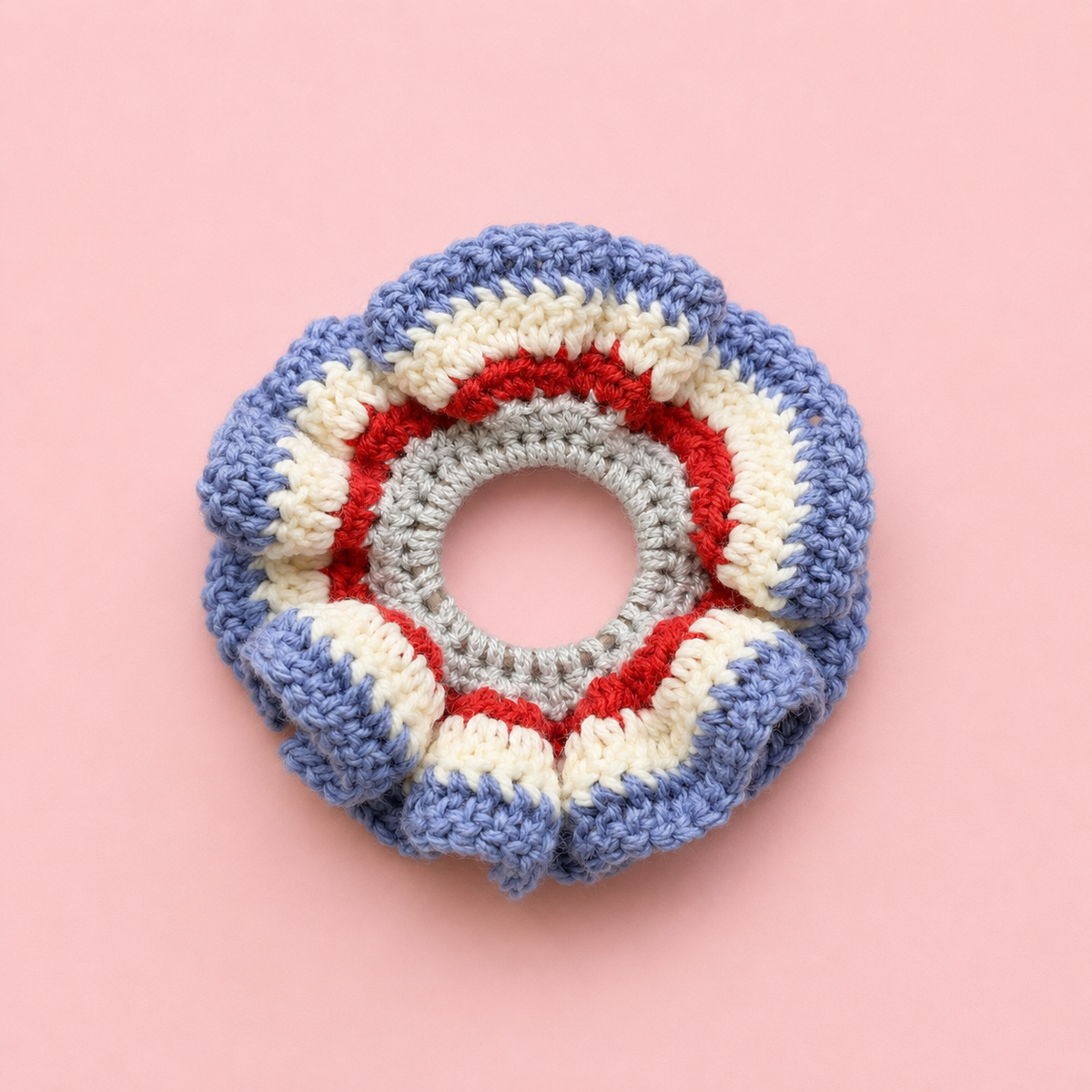 One of a kind handmade crochet scrunchie in blue, cream, red and grey stripes on a pale pink background.
