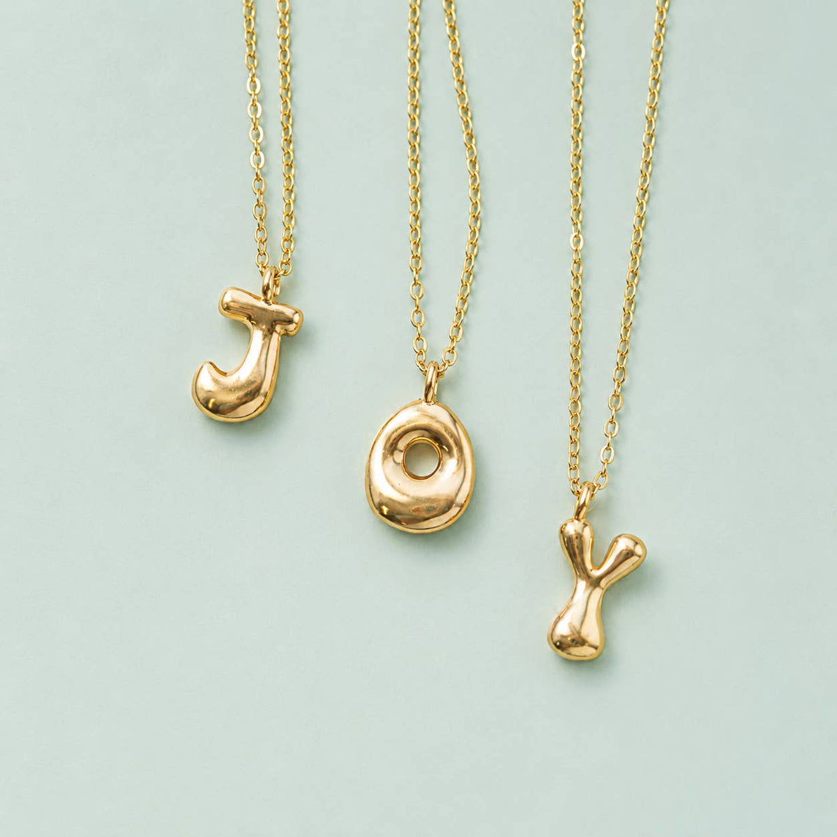 Gold J, O and Y bubble initial necklaces on delicate gold chains displayed against a pale blue background.