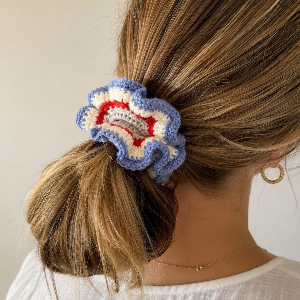A close-up lifestyle shot of a woman from the back, showing a hand-crocheted ruffled hair scrunchie in blue, cream, and red holding her light brown hair in a low ponytail.