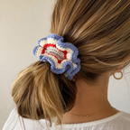 A close-up lifestyle shot of a woman from the back, showing a hand-crocheted ruffled hair scrunchie in blue, cream, and red holding her light brown hair in a low ponytail.