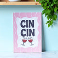 Cheerful Cin Cin A5 art print with bold navy lettering, clinking wine glasses and a wavy pink border displayed in a bright kitchen.