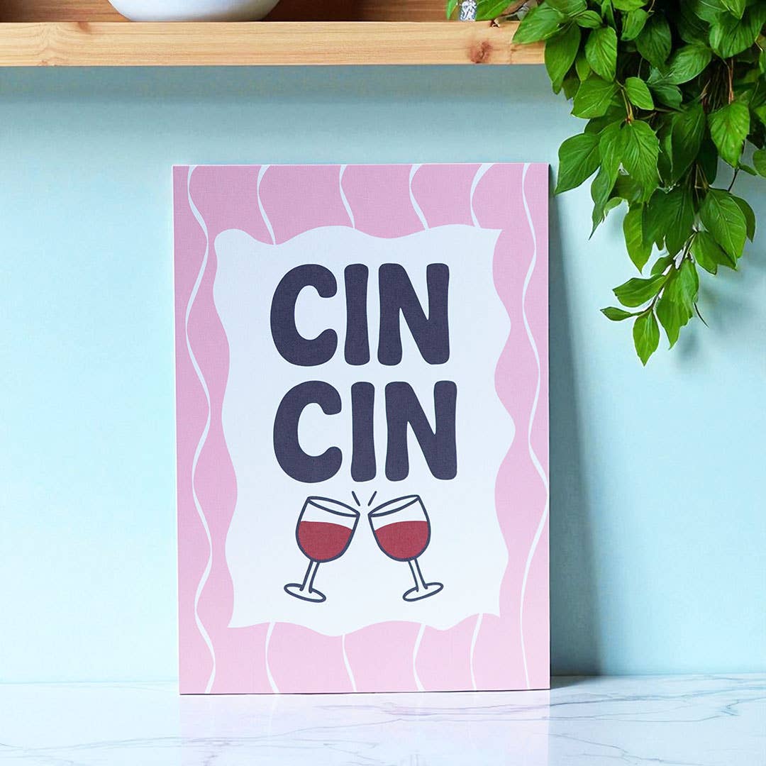 Cheerful Cin Cin A5 art print with bold navy lettering, clinking wine glasses and a wavy pink border displayed in a bright kitchen.