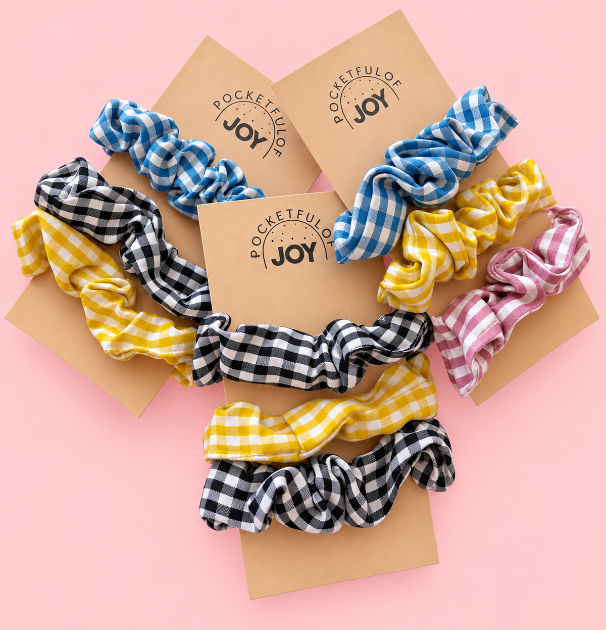Set of colourful gingham scrunchies in blue, yellow, pink and black and white checks, displayed on Pocketful of Joy cards against a pink background.