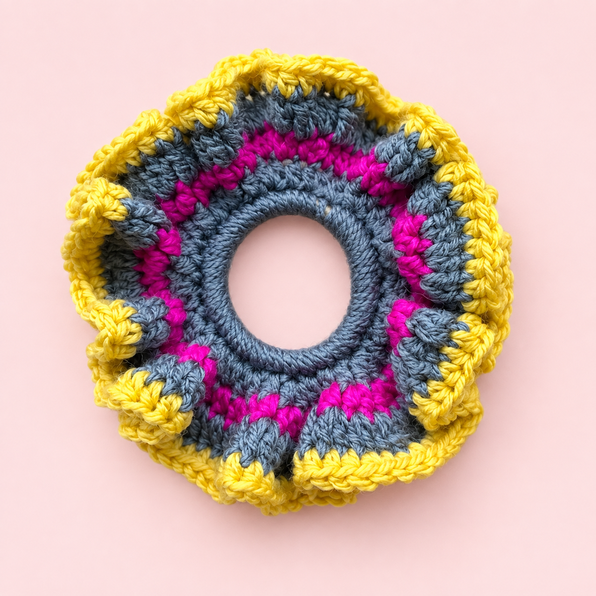 Handmade crochet scrunchie in yellow, grey and hot pink with a ruffled design on a pale pink background.