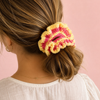 A close-up lifestyle shot of a woman from behind, wearing a handmade crocheted scrunchie in yellow and pink on her brown hair, set against a solid pink background.