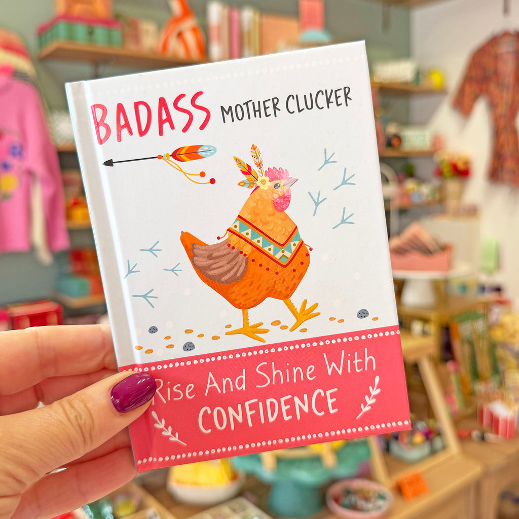 A hand with purple nail polish holds a small notebook titled 'BADASS MOTHER CLUCKER' in a colorful shop setting. The notebook cover features an illustration of a decorated orange hen and a pink banner at the bottom with the text 'Rise And Shine With CONFIDENCE'.