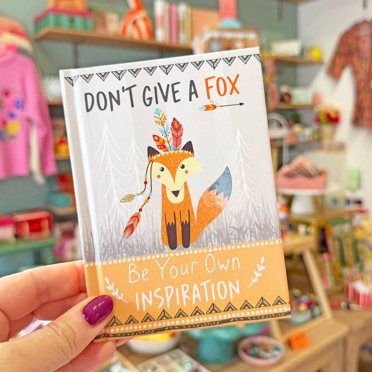A hand with purple nail polish holds a small notebook or journal featuring a stylized fox illustration and the text 'DON'T GIVE A FOX Be Your Own INSPIRATION'. The background shows a softly blurred craft or stationery store interior with shelves and colorful items.