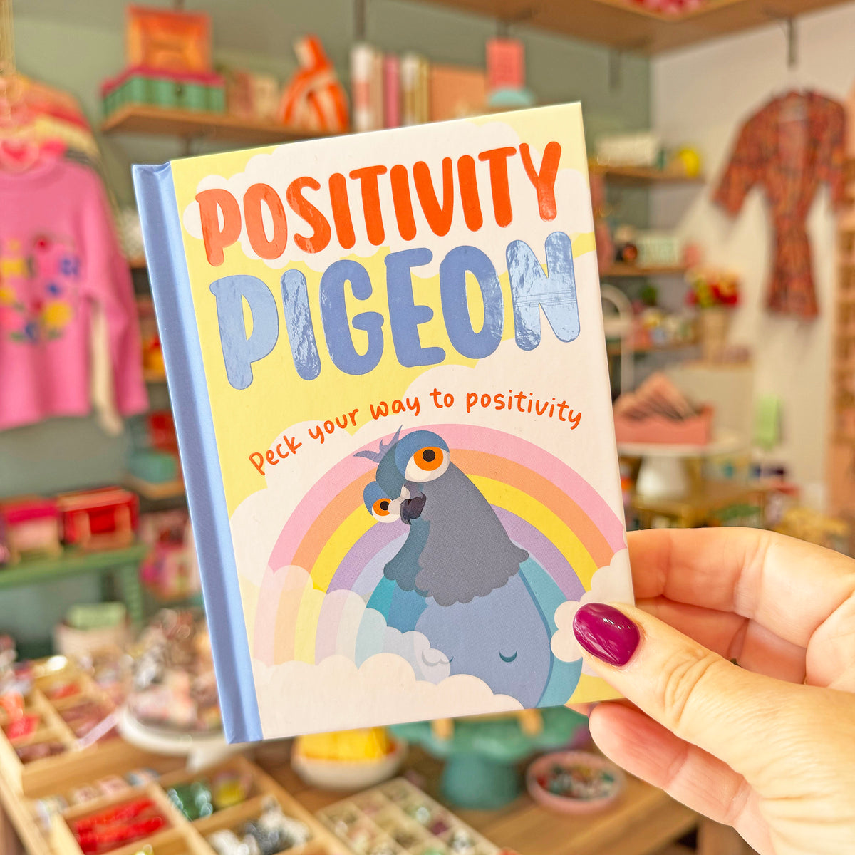 A hand with purple nail polish holds a small hardcover book titled 'POSITIVITY PIGEON' in a colorful shop setting. The book cover features a blue pigeon under a rainbow on a pale yellow background.