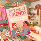A hand with purple nail polish holds a pink hardcover book titled 'WHY WE'RE FRIENDS .