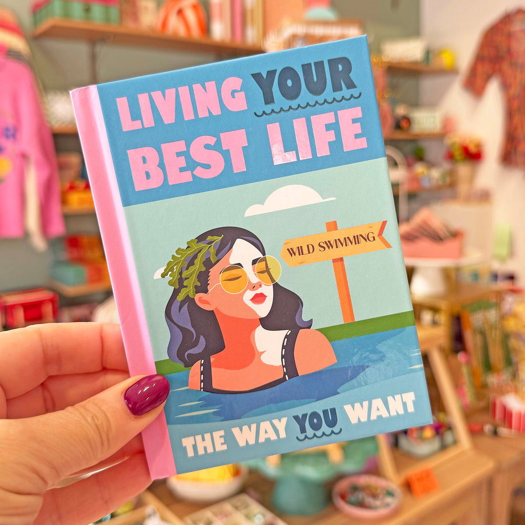 A hand with purple nail polish holds a colorful hardcover book titled 'LIVING YOUR BEST LIFE' in a blurred retail store setting.