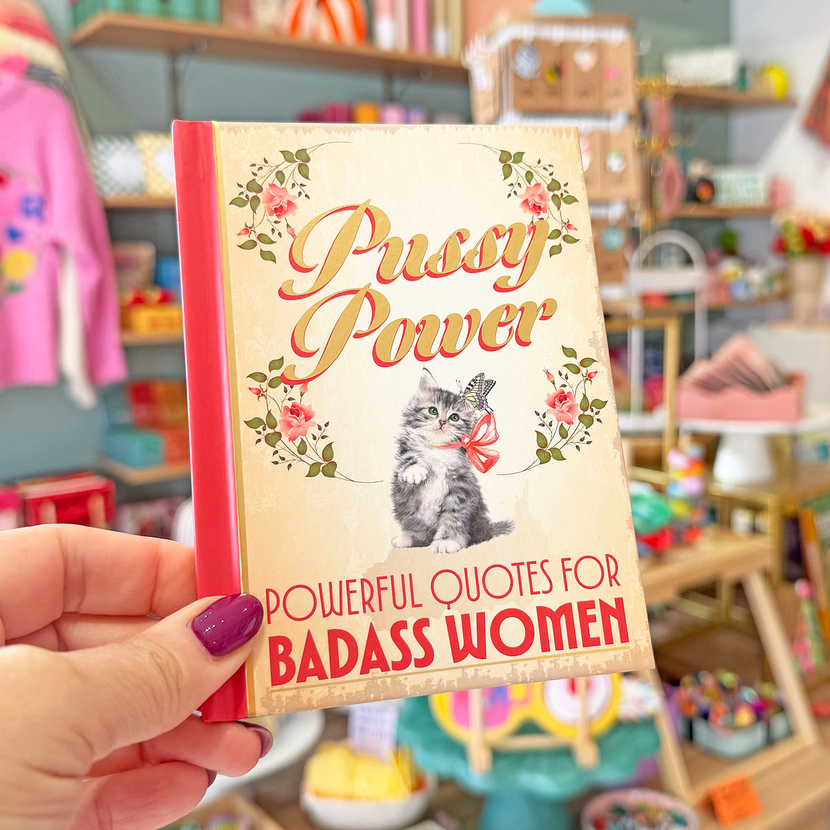 A hand with purple nail polish holds a small book titled 'Pussy Power' in a colorful, blurred boutique setting. The book cover features a vintage-style kitten illustration and floral accents.