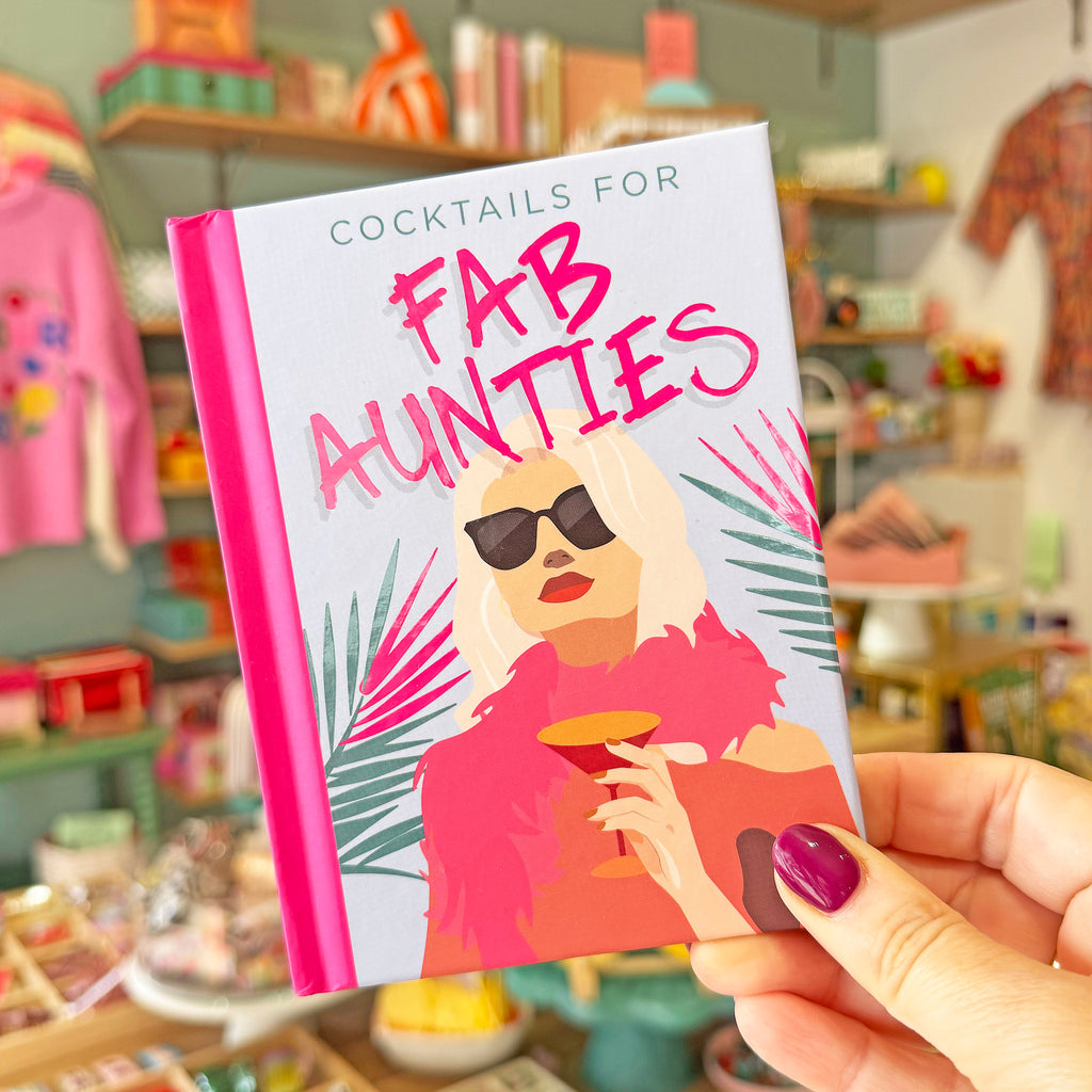 A hand with purple nail polish holds a small cocktail-themed book or notebook titled 'COCKTAILS FOR FAB AUNTIES' against a blurred shop background.
