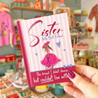 A hand with purple nail polish holds a small pink and white striped book or notebook titled 'Sister My Best Friend' in a colorful, blurred boutique setting.