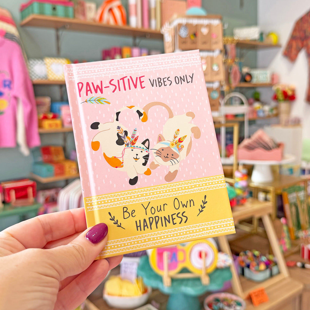 A hand with purple nail polish holds a pink notebook titled 'PAW-SITIVE VIBES ONLY' in a colorful, blurred boutique setting. The notebook features cartoon cats, a yellow band reading 'Be Your Own HAPPINESS', and a pink polka-dot cover.