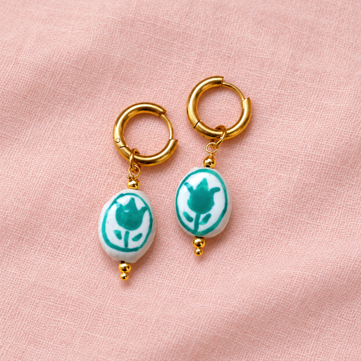 A pair of gold hoop earrings with oval white ceramic pendants featuring a teal tulip design, displayed on a soft pink textured fabric background.