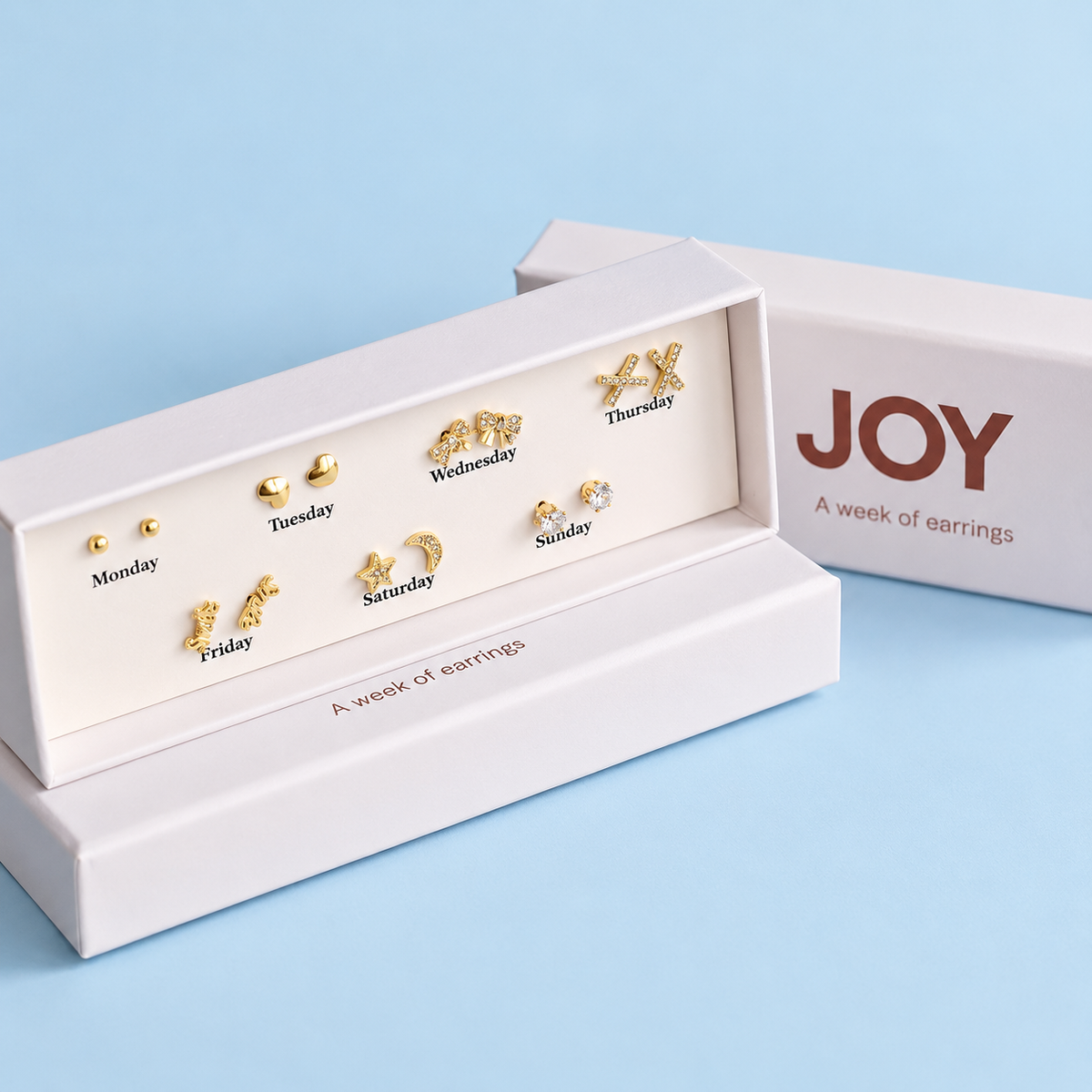 Open JOY Week of Earrings gift set showing seven pairs of gold plated stud earrings against a pale blue background.