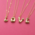 Gold bubble initial necklaces spelling LOVE on delicate gold chains against a bright pink background.