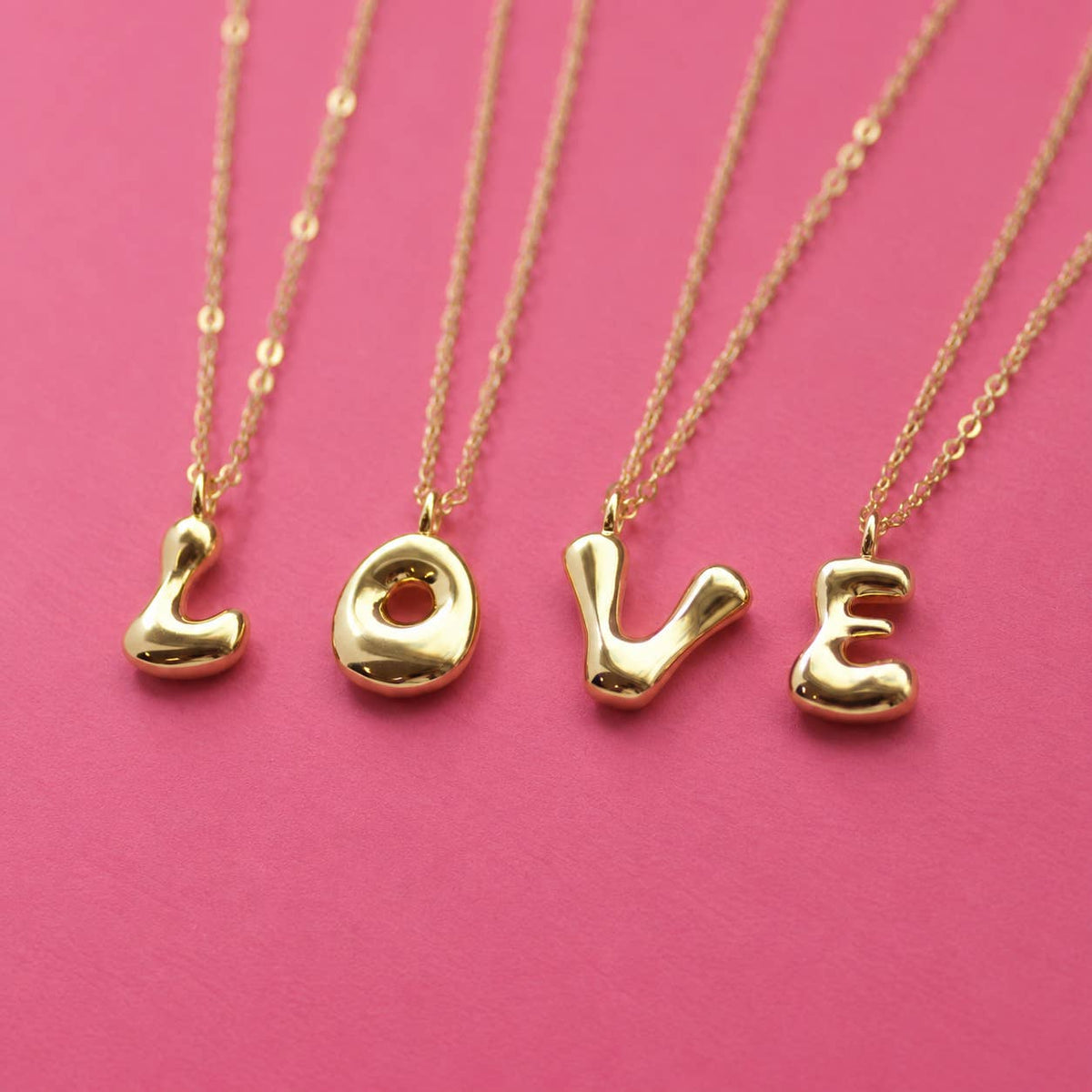 Gold bubble initial necklaces spelling LOVE on delicate gold chains against a bright pink background.