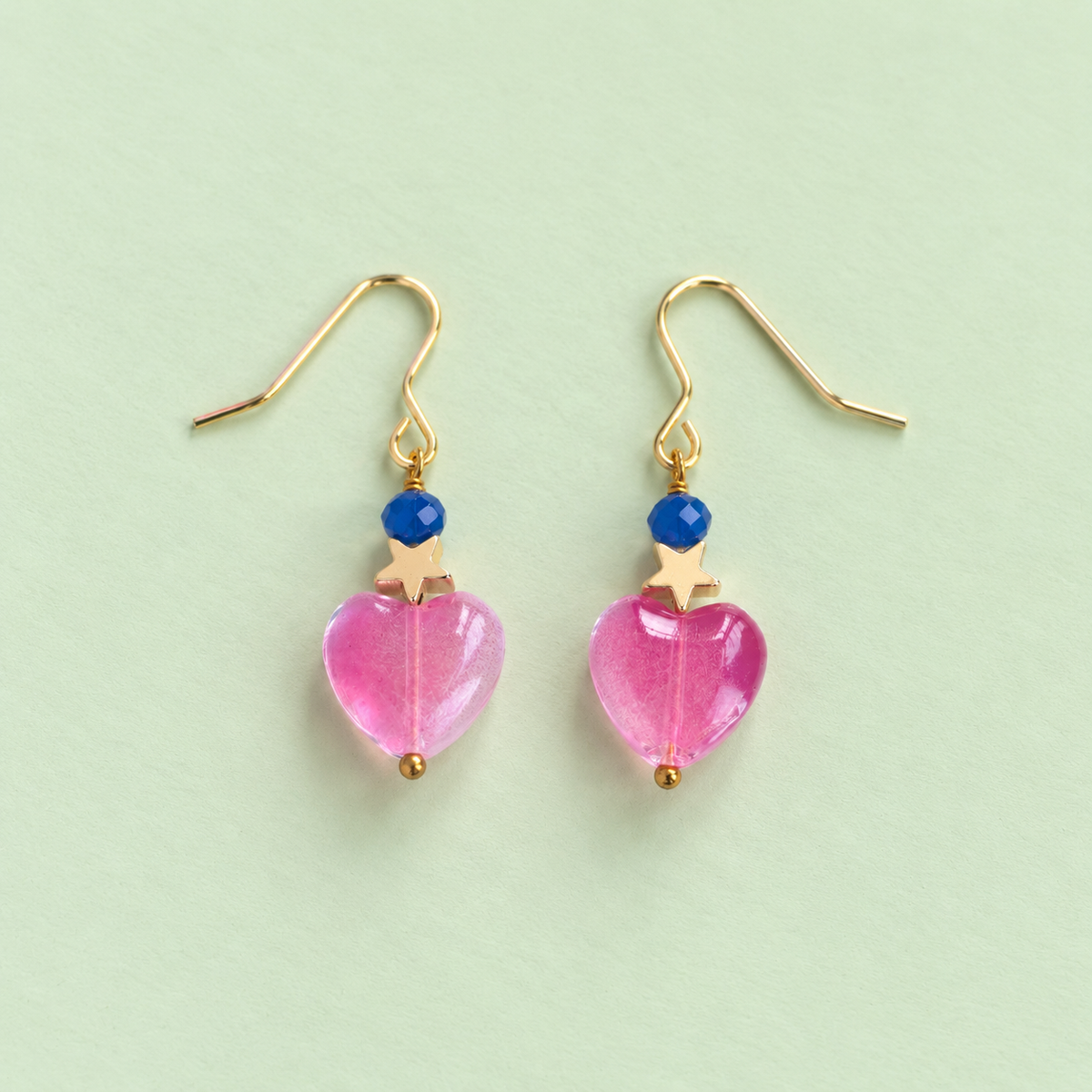 One of a kind gold plated drop earrings with bright pink glass heart beads, blue faceted beads and small gold star details on a pale mint background.