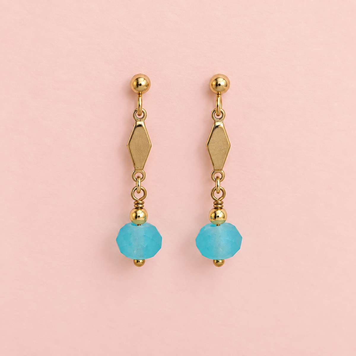 Pair of one of a kind gold plated drop earrings with geometric details and frosted aqua blue faceted glass beads on a pale pink background.